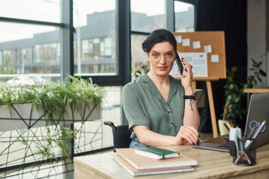 Düşünceli, güzel, engelli, gözlüklü, iş yerinde telefonla konuşan bir kadın.