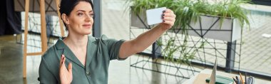 Çekici iş kadını, günlük kıyafetleriyle, video ve afiş sırasında telefon kamerasına el sallıyor.