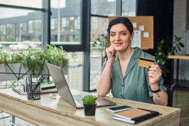 attractive jolly disabled businesswoman in wheelchair holding credit card and looking at camera clipart