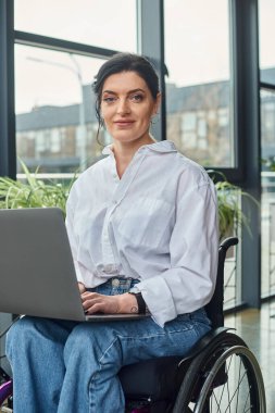 Tekerlekli sandalyedeki engelli neşeli iş kadını laptopuyla çalışırken kameraya bakıyor.