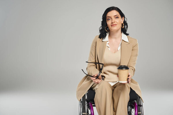 привлекательная женщина-инвалид в пастельном костюме держит солнцезащитные очки и кофе и смотрит в камеру