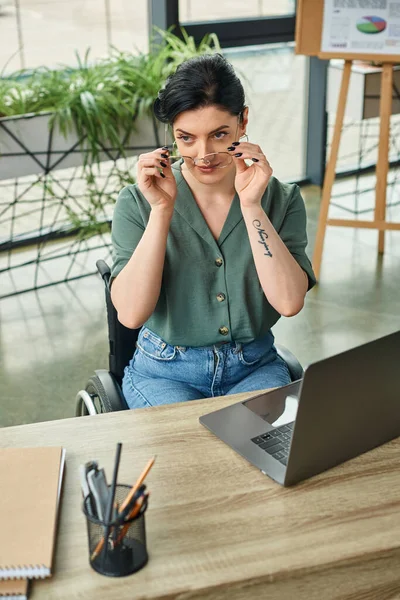 attractive businesswoman with glasses in casual attire in wheelchair working at her laptop in office