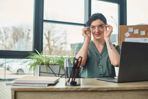 beautiful disabled woman in casual attire with tattoo and glasses looking away while in office