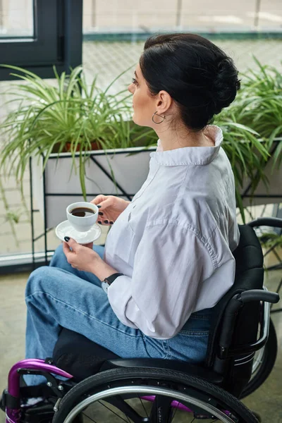 attractive dedicated disabled woman in stylish attire sitting in wheelchair with coffee cup