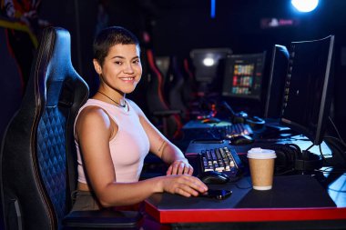 Mutlu zoomer yaşında bir kadın bilgisayar monitörü, kahve ve siber spor oyunlarıyla bir masada oturuyor.