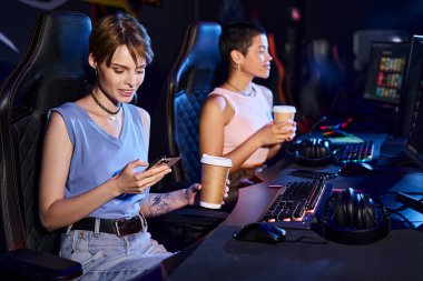 Bilgisayar masasında oturan bir kadın elinde telefon ve kahveyle bilgisayar sporları kulübünde arkadaşının yanında.