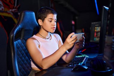 Odaklanmış bir kadın telefonuyla dijital dünyaya dalarken bilgisayarının başına rahatça oturur.