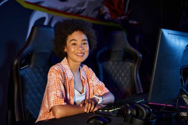 Gülümseyen Afro-Amerikan kadın, elinde akıllı bir el kamerasıyla bilgisayarın yanındaki siber spora bakıyor.