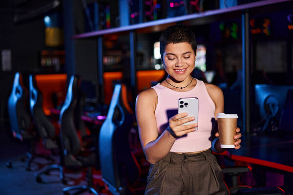 stylish woman, clad in fashionable clothing, smiles as she holds her phone and a cup of coffee