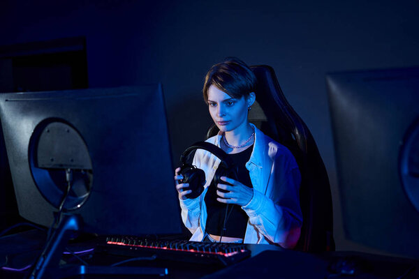 сфокусированная женщина, держащая наушники и глядя на компьютер в синей комнате, концепция киберспорта