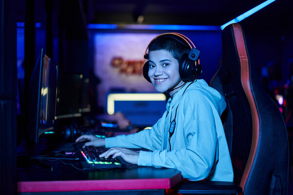 cheerful short haired woman looking at camera near computer monitor in blue lit room, zoomer