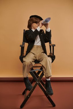 Şık giyinmiş yakışıklı bir çocuk yönetmen koltuğunda oturur ve rulo kağıttaki delikten bakar.