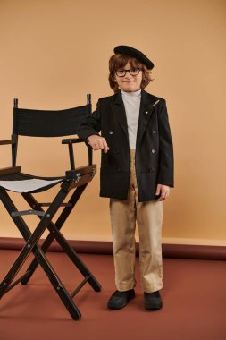 happy boy stands proudly next to director chair, wearing coat and trousers, with a big smile on face clipart