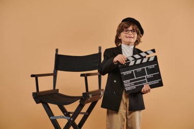 young filmmaker in trendy clothing, happily poses with a clapperboard in hand against beige wall clipart