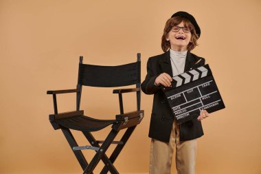 excited filmmaker boy in trendy clothing happily poses with clapperboard in hand against beige wall clipart