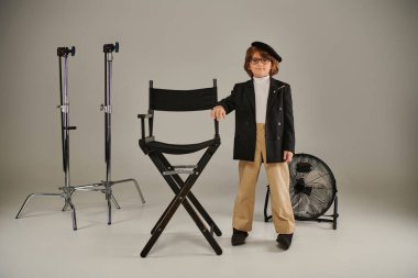 cute boy in beret and smart casual attire stands confidently near director chair on grey backdrop clipart