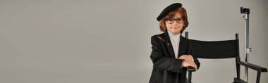 cute boy in beret and stylish attire stands confidently near director chair on grey backdrop, banner clipart