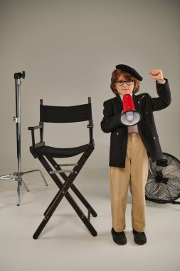A young activist in beret stands with megaphone and director chair on grey, boy as filmmaker clipart