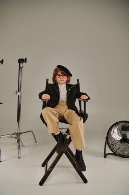 confident filmmaker boy in beret and glasses sitting on director chair on grey backdrop clipart