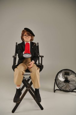 confident filmmaker boy in beret and glasses sitting on director chair and holding red megaphone clipart