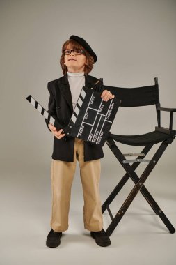 Şık bir yönetmen, bereli, elinde alkış tahtası olan ve yönetmen koltuğunun yanında duran gözlüklü çocuk.