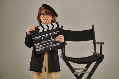 Şık bir film yapımcısı, bereli, elinde alkış tahtası ve yönetmen koltuğunun yanında duran gözlüklü çocuk.