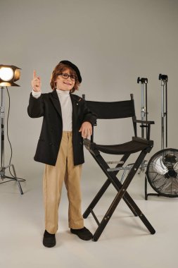 happy stylish boy in beret and glasses standing in front of director chair, softbot on background clipart