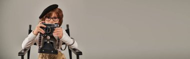 Beredeki çocuk, yönetmen koltuğunda, fotoğrafçı pankartıyla otururken eski model kamerayla fotoğraf çekiyor.
