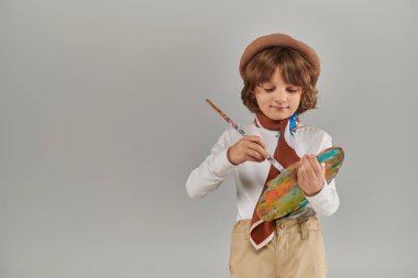 Boyalı fırça ve palet tutan genç bir ressam, gri arka planda duran bereli ve eşarplı şirin bir çocuk.
