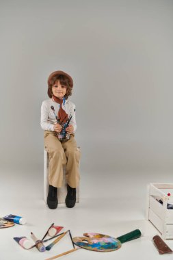 Genç sanatçı elinde cam kavanozla gri renkte boya fırçaları, bereli çocuk ve sandalyede oturan eşarp