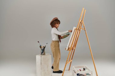 Ahşap sehpanın yanında paletle duran genç ressam, bereli ve eşarplı şık çocuk.