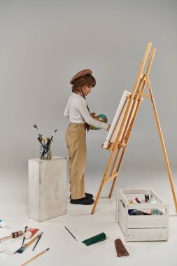 Şövalenin yanında palet ve boya fırçası olan genç bir sanatçı, bereli ve eşarplı şık bir çocuk.