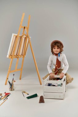 Mutlu genç sanatçı tahta kutuda boyayı seçiyor, bereli çocuk sehpanın yanında oturuyor ve fırça boyuyor.
