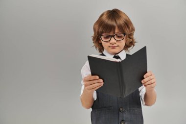 Gözlüklü meraklı çocuk, gri arka plan üzerine bir kitap okurken hayal dünyasının derinliklerine iniyor.
