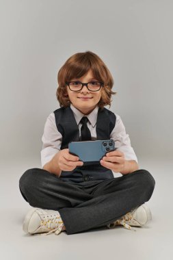 Gözlüklü ve şık giyinmiş gülümseyen çocuk gri üzerinde mobil oyun oynuyor, akıllı telefon tutuyor.