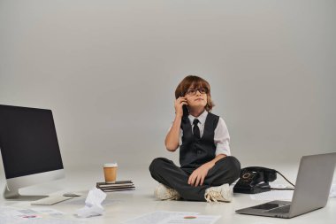 Gözlüklü ve resmi kıyafetli bir çocuk akıllı telefondan konuşuyor ve ofis ekipmanlarıyla çevrili oturuyor.