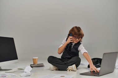 Gözlüklü ve resmi giyinen çocuk akıllı telefondan konuşuyor ve ofis ekipmanlarıyla çevrili oturuyor.