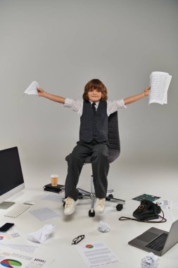 Ofis malzemeleriyle çevrili, elinde kağıtlarla sandalyede oturan mutlu çocuk.