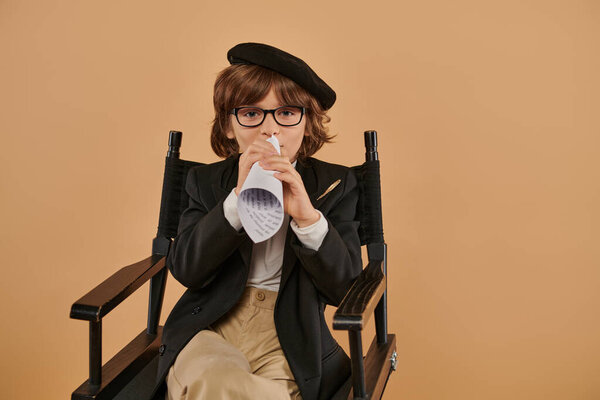 stylish boy in beret and glasses sits in director chair, speaking in piece of rolled paper