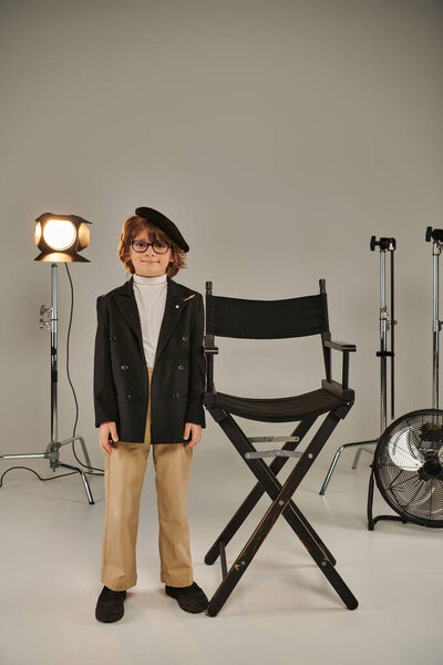 Хорошо одетый мальчик в берцах и очках, стоящий перед режиссерским креслом, трезвый на заднем плане