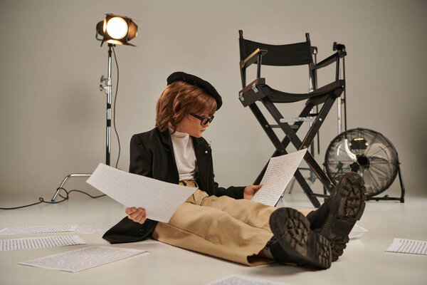 child in glasses and beret reading screenplay and sitting on floor, boy as director of filmmaker