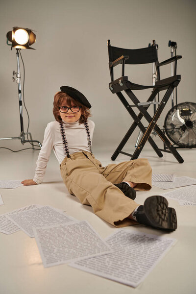 boy in suspenders and beret sitting on floor surrounded by screenplay, boy as director of filmmaker