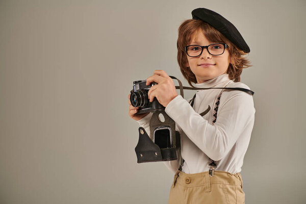pleased boy, young photographer in beret and suspenders taking photo on retro camera in studio