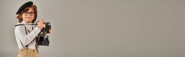 happy young photographer in beret and suspenders taking photo on retro camera in studio, banner