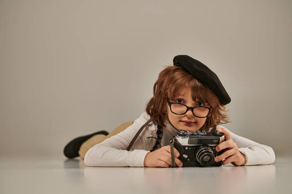 happy young photographer in beret and suspenders taking photo on retro camera and lying on floor