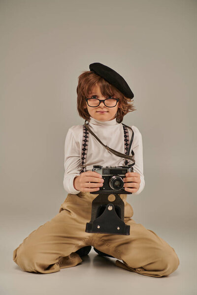 young photographer in beret and glasses holding camera and sitting on floor, boy in suspenders