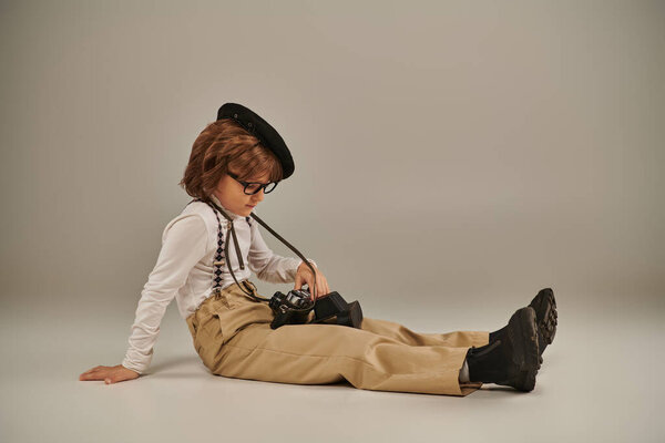 young photographer in beret and glasses holding camera and sitting on floor, cute boy in suspenders