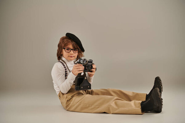 young photographer in beret and glasses holding camera and sitting on floor, cute kid in suspenders
