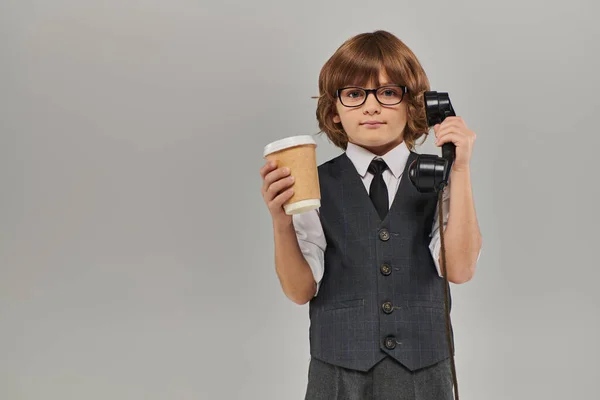 elegant boy in glasses and formal attire holding retro phone and drink in paper cup on grey