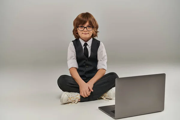 stock image boy in elegant attire with vest and trousers sitting near laptop on grey, future businessman